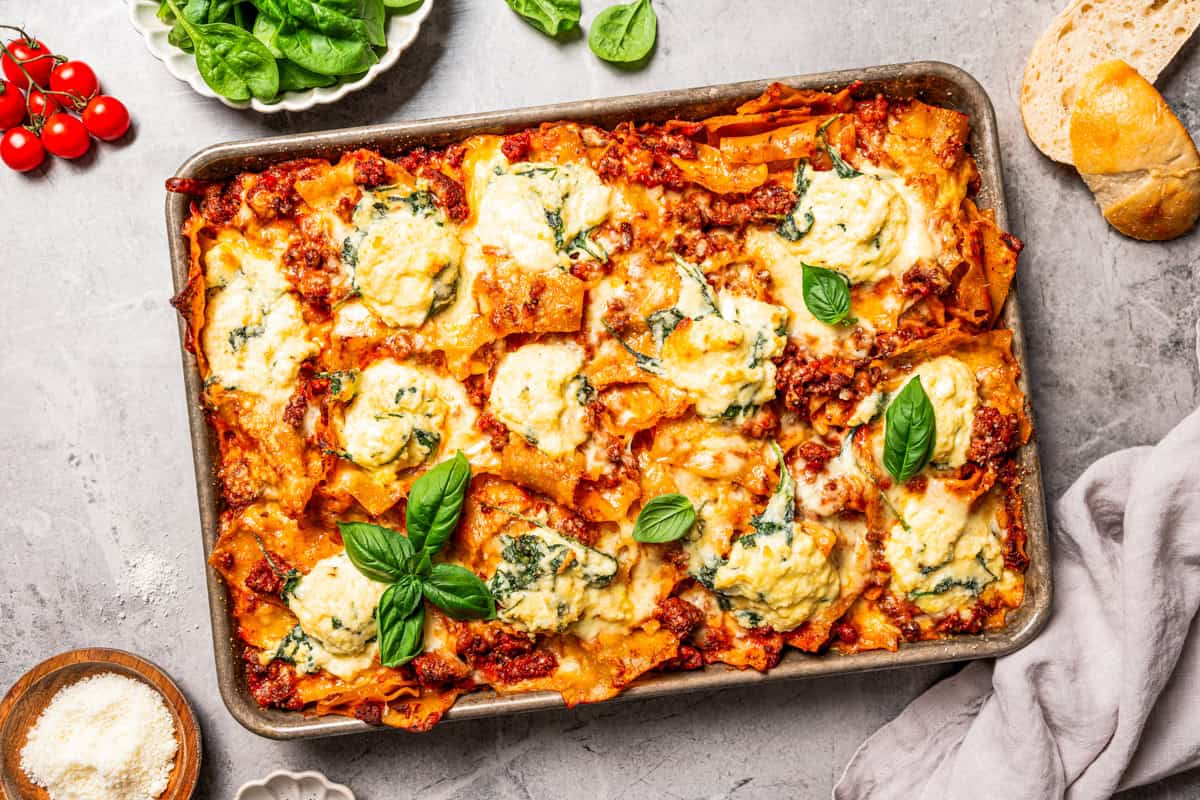 Sheet pan lasagna is a cheesy, delicious, golden-baked lasagna recipe with crispy edges and no layering required. Mix, dump, and bake! Overhead view of a cheesy sheet pan lasagna garnished with fresh basil leaves.