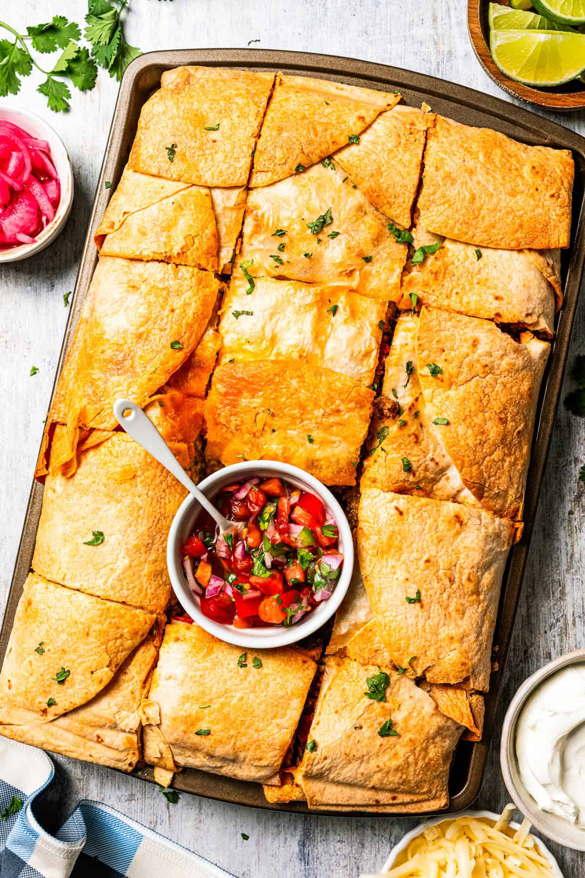 Baked sheet pan tacos are an easy way to make tacos for a crowd with crispy tortillas, seasoned ground beef, melty cheese, and pico de gallo. A sheet pan of baked tacos with a bowl of homemade pico de gallo placed in the center.