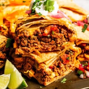 Stacked sheet pan tacos with crispy tortillas, filled with ground beef, melted cheese, and vegetables, topped with sour cream and fresh cilantro.