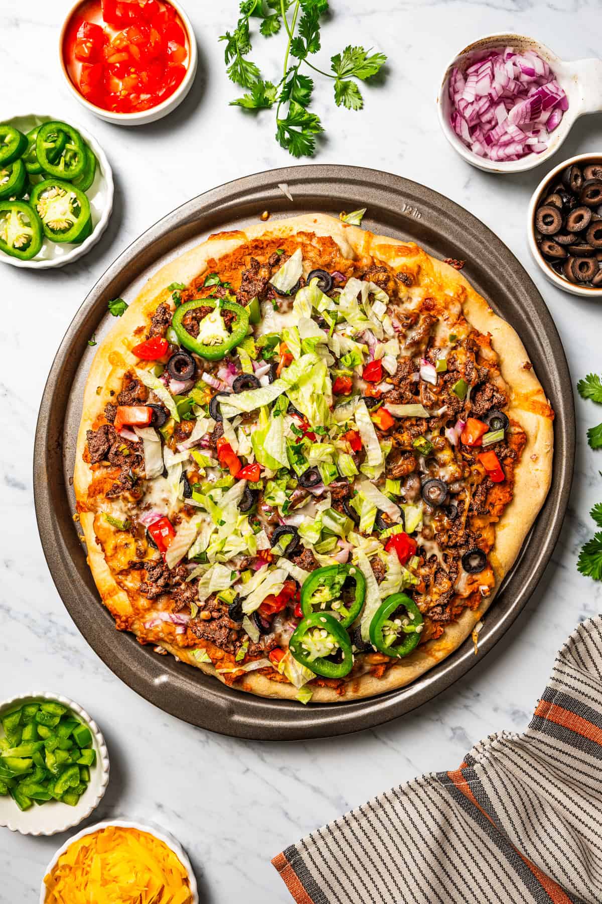 This Mexican-inspired taco pizza recipe has a crispy pizza crust topped with refried beans, taco meat, salsa, and your favorite taco toppings. A pan of pizza garnished with taco toppings.