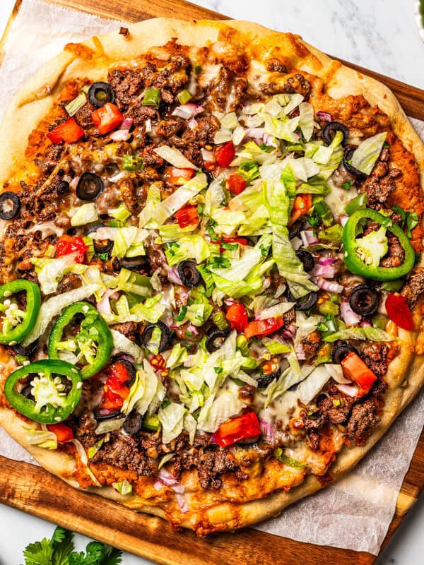 Taco pizza garnished with sour cream, jalape&ntilde;os, and taco toppings on a wooden cutting board.