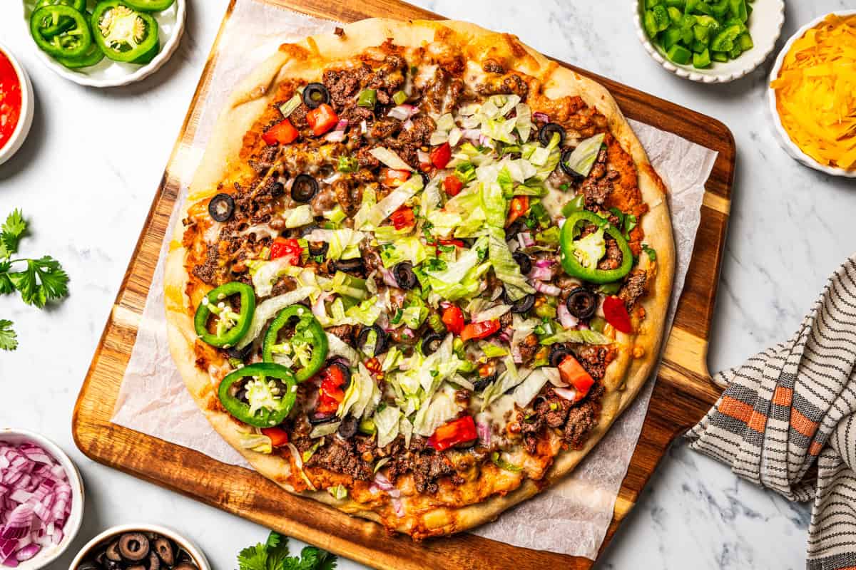 This Mexican-inspired taco pizza recipe has a crispy pizza crust topped with refried beans, taco meat, salsa, and your favorite taco toppings. Taco pizza garnished with sour cream, jalapeños, and taco toppings on a wooden cutting board.