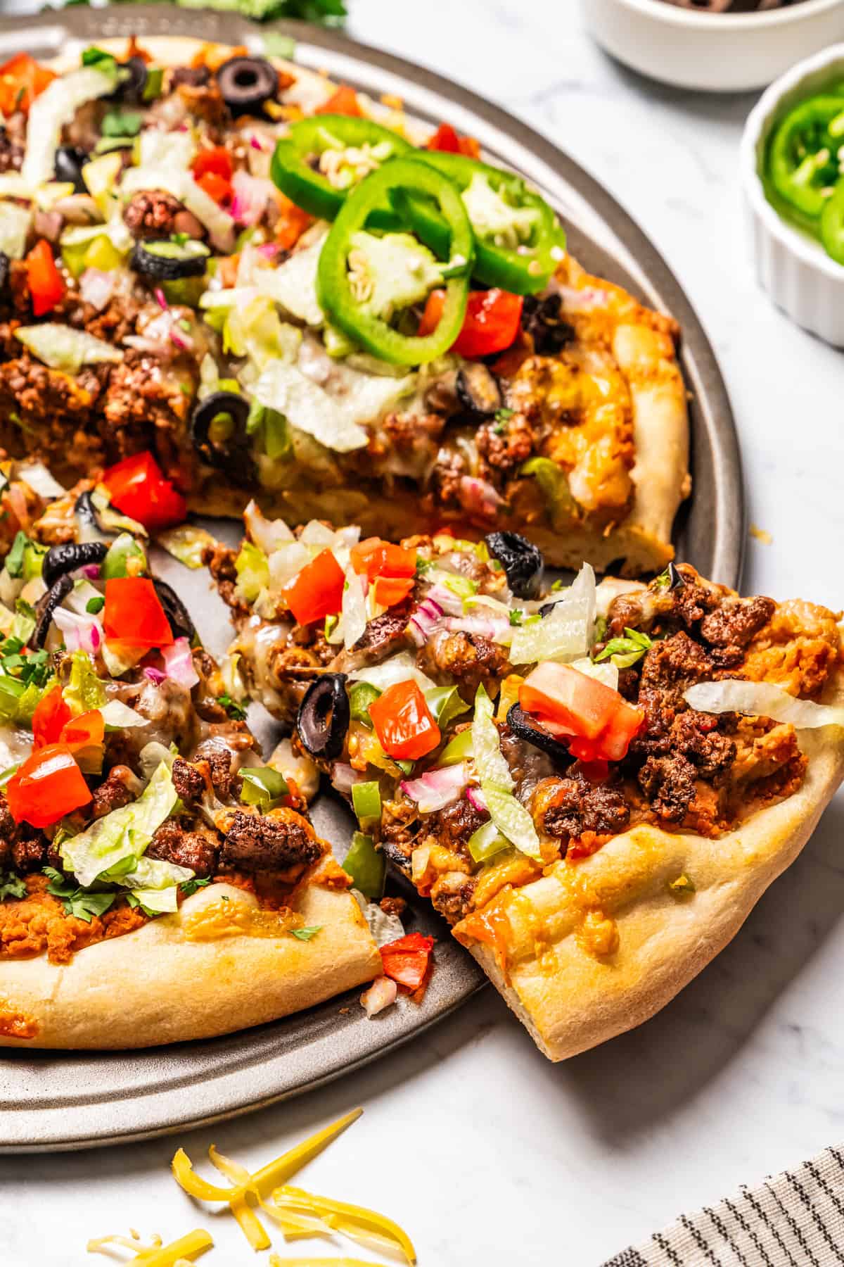 This Mexican-inspired taco pizza recipe has a crispy pizza crust topped with refried beans, taco meat, salsa, and your favorite taco toppings. A slice of taco pizza partially removed from the rest of the pizza on a pan.