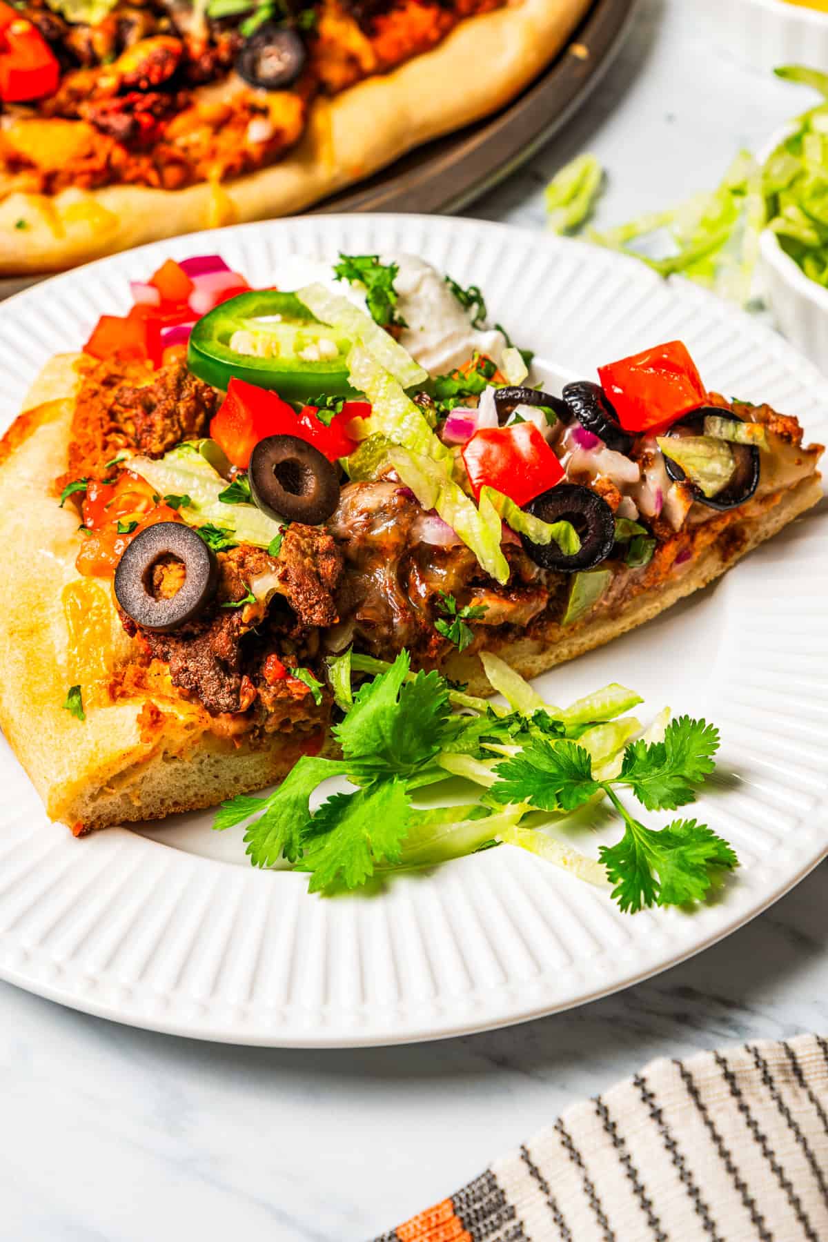 This Mexican-inspired taco pizza recipe has a crispy pizza crust topped with refried beans, taco meat, salsa, and your favorite taco toppings. A slice of taco pizza served on a dinner plate.