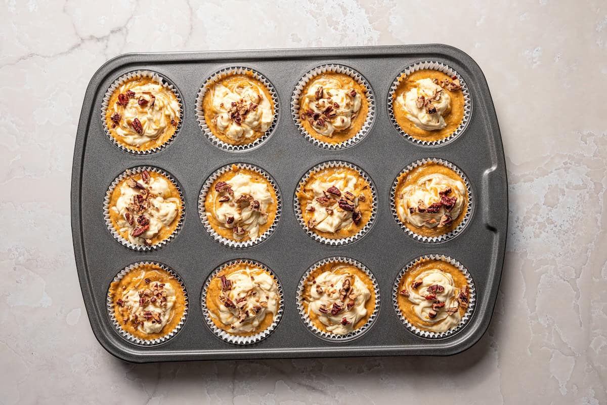 These pumpkin cream cheese muffins are moist, spiced pumpkin muffins swirled with cream cheese filling and topped with toasted pecans. Pumpkin cream cheese muffins batter topped with chopped pecans, ready to bake in a muffin pan.