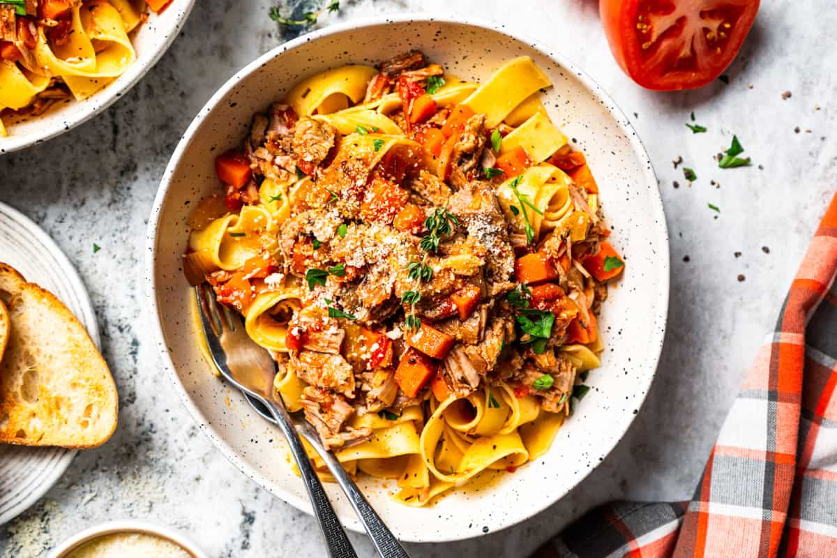 This slow-braised pork ragu recipe cooks for hours in the oven until fall-apart tender and ready to shred into a rich, flavorful meat sauce. A bowl of pork ragu with pappardelle pasta served with toasted bread on the side and garnished with Parmesan cheese.