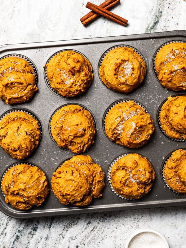 Protein pumpkin muffins baked in a muffin tin with pumpkin seeds, cinnamon, and maple syrup nearby.
