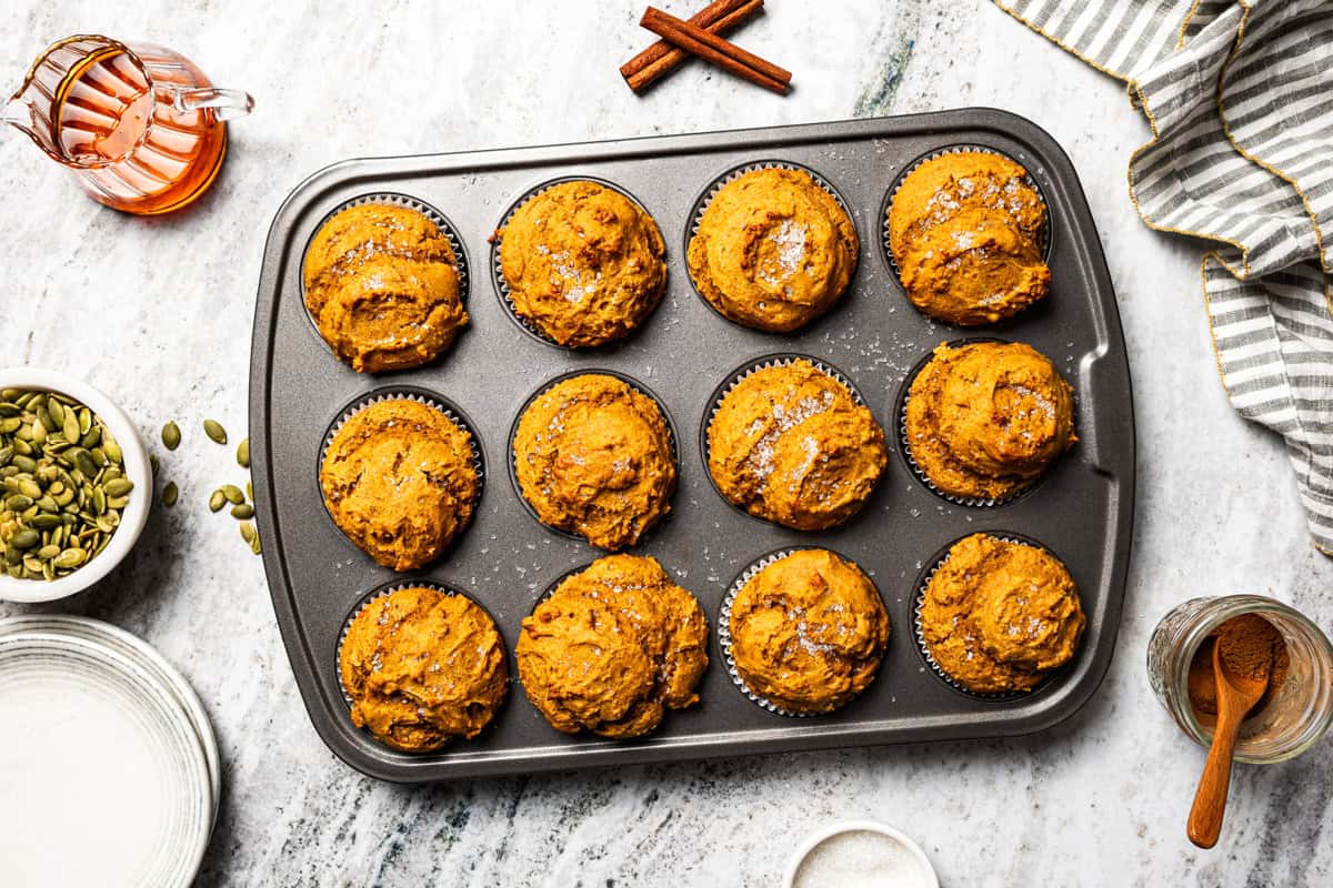 Moist, fluffy pumpkin protein muffins are a high-protein fall treat made with vanilla whey powder, Greek yogurt, eggs, and pumpkin purée. Protein pumpkin muffins baked in a muffin tin with pumpkin seeds, cinnamon, and maple syrup nearby.