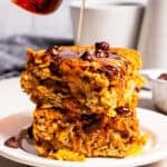 Pouring maple syrup over two slices of pumpkin baked oatmeal stacked on a plate.