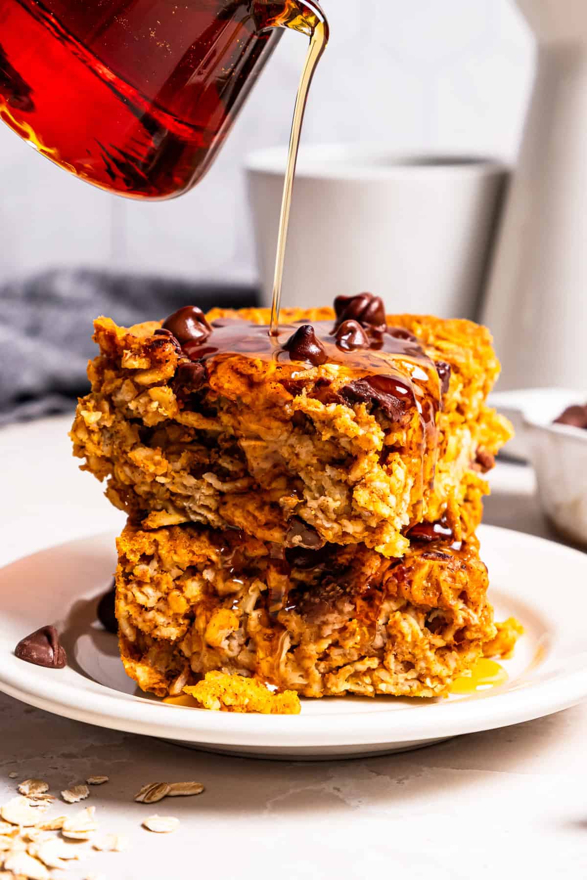 Pumpkin baked oatmeal is the perfect easy, make-ahead fall breakfast! Filled with hearty oats, warm spices, pumpkin purée, and maple syrup. Pouring maple syrup over two slices of pumpkin baked oatmeal stacked on a plate.