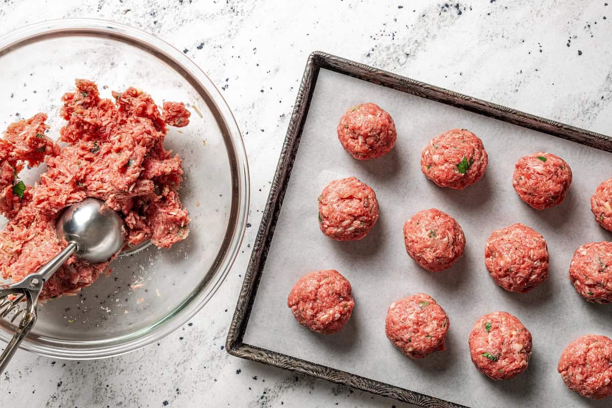 Not your old-fashioned porcupine meatballs! Make these with juicy ground beef, sausage, rice, and herbs, cooked in homemade tomato sauce. Rolled porcupine meatballs arranged on a baking sheet next to meatball mixture in a bowl.