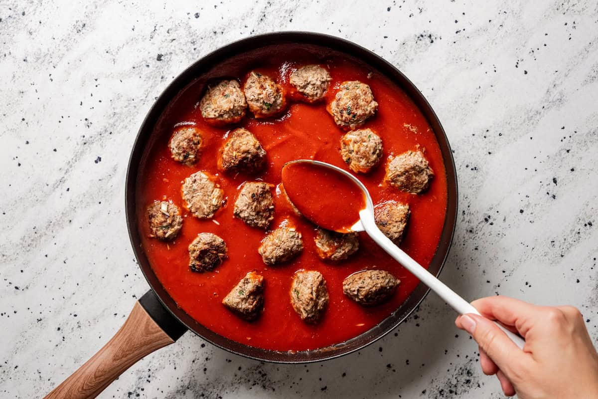 Not your old-fashioned porcupine meatballs! Make these with juicy ground beef, sausage, rice, and herbs, cooked in homemade tomato sauce. Spooning garlic parmesan tomato sauce over porcupine meatballs in a skillet.