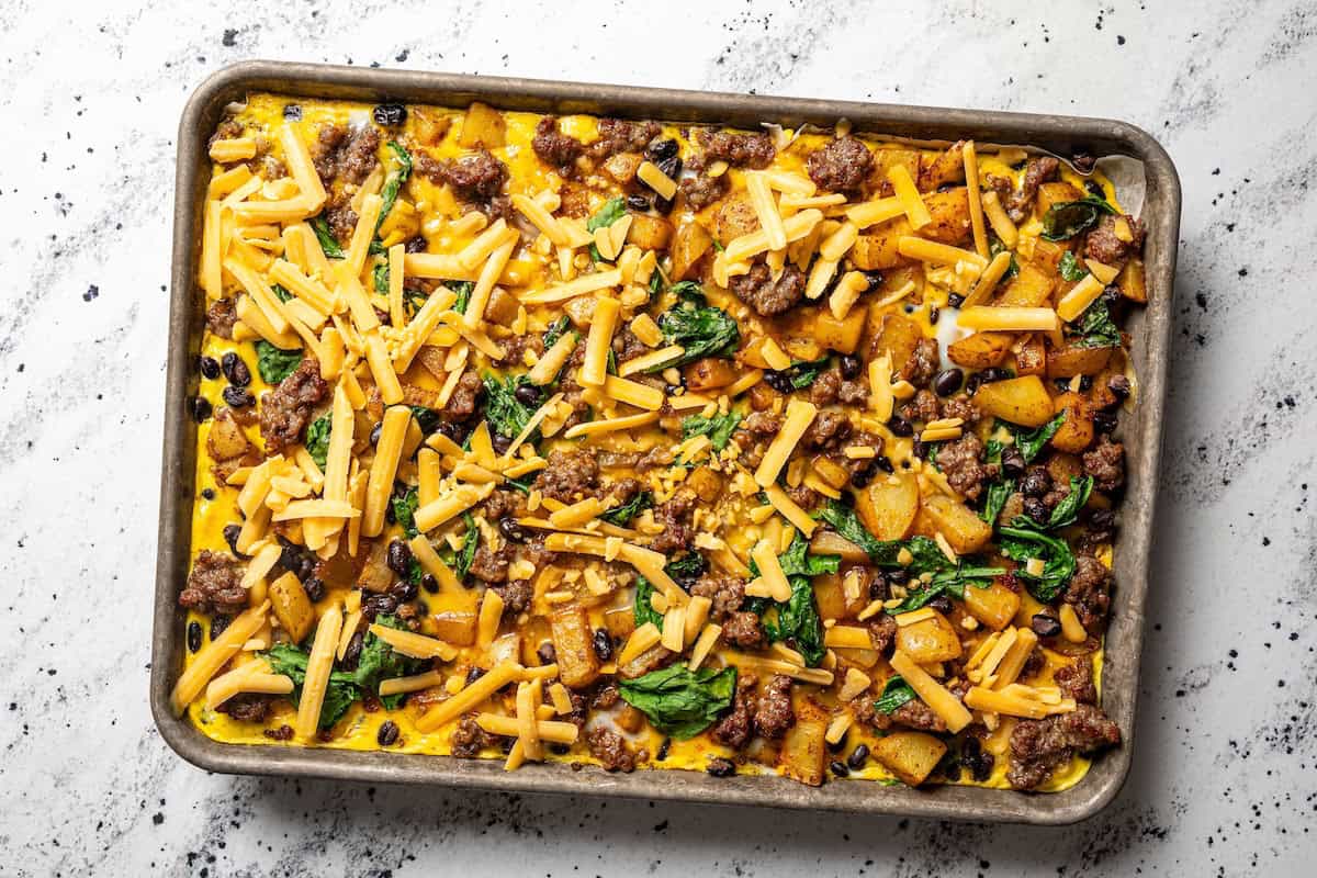 Sheet pan breakfast burritos are an easy way to batch-make breakfast burritos! Bake the filling in one pan, then roll it into soft tortillas. Shredded cheese sprinkled over sheet pan breakfast burritos ingredients on a baking pan.