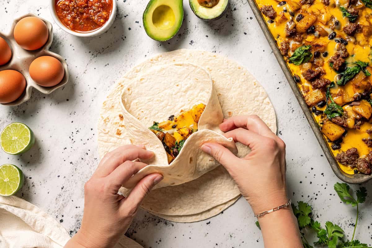 Sheet pan breakfast burritos are an easy way to batch-make breakfast burritos! Bake the filling in one pan, then roll it into soft tortillas. Two hands folding a flour tortilla around the sheet pan breakfast burrito filling.