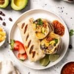 Overhead view of sheet pan breakfast burritos on a plate, with one burrito cut in half to show the egg and sausage filling.