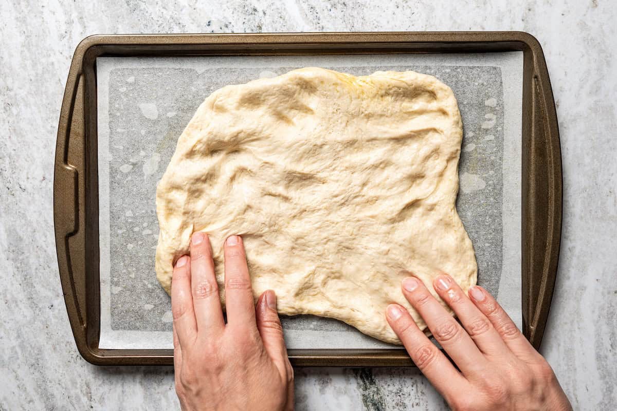 Make this golden, crispy sheet pan pizza recipe with easy homemade dough and your favorite flavorful toppings, like pepperoni and cheese. Two hands stretching homemade pizza dough over a lined sheet pan.