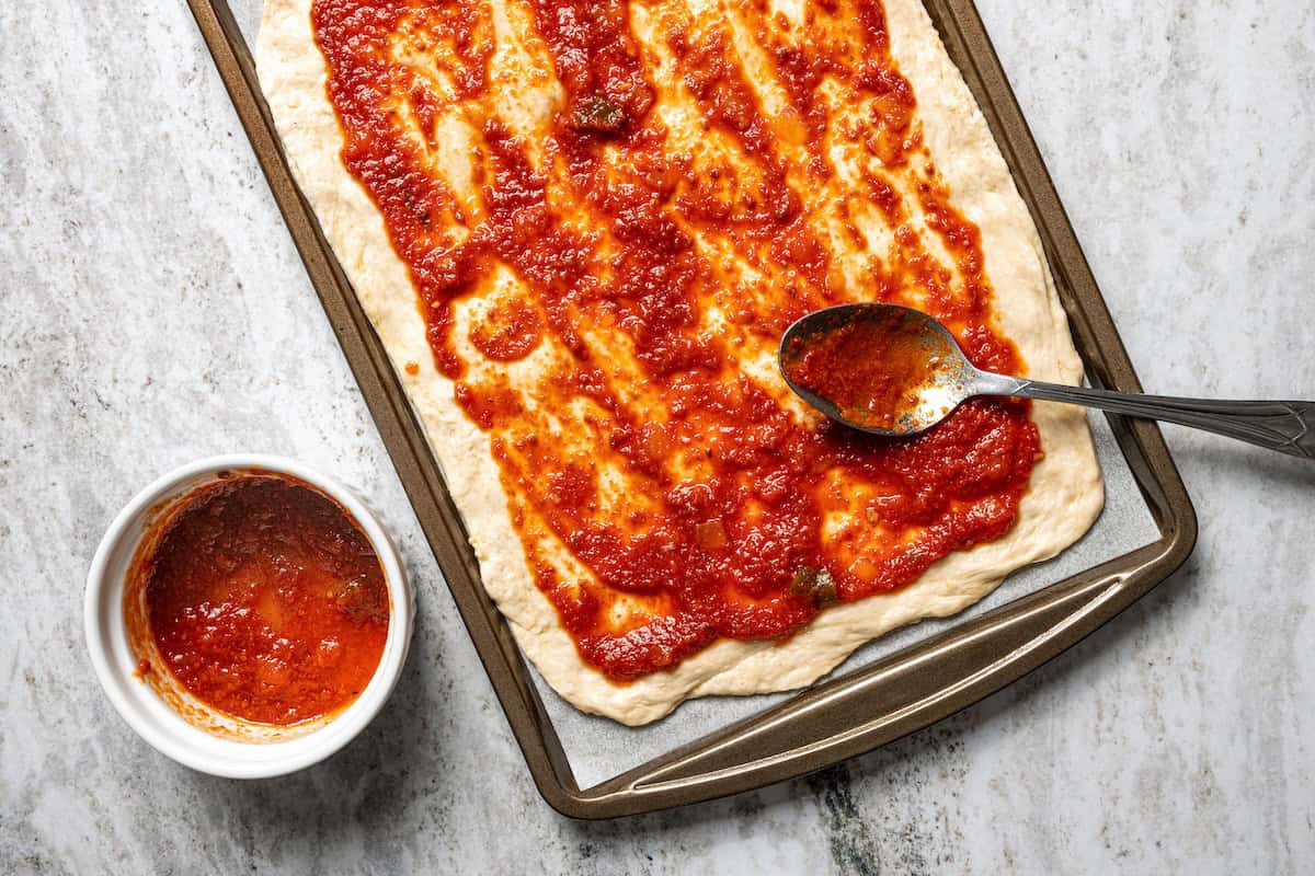 Make this golden, crispy sheet pan pizza recipe with easy homemade dough and your favorite flavorful toppings, like pepperoni and cheese. A spoon spreading pizza sauce over a sheet pan pizza crust.