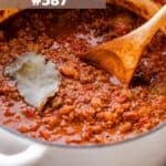 A pot of no-bean chili with ground beef, tomatoes, a bay leaf, and a wooden spoon stirring through it.