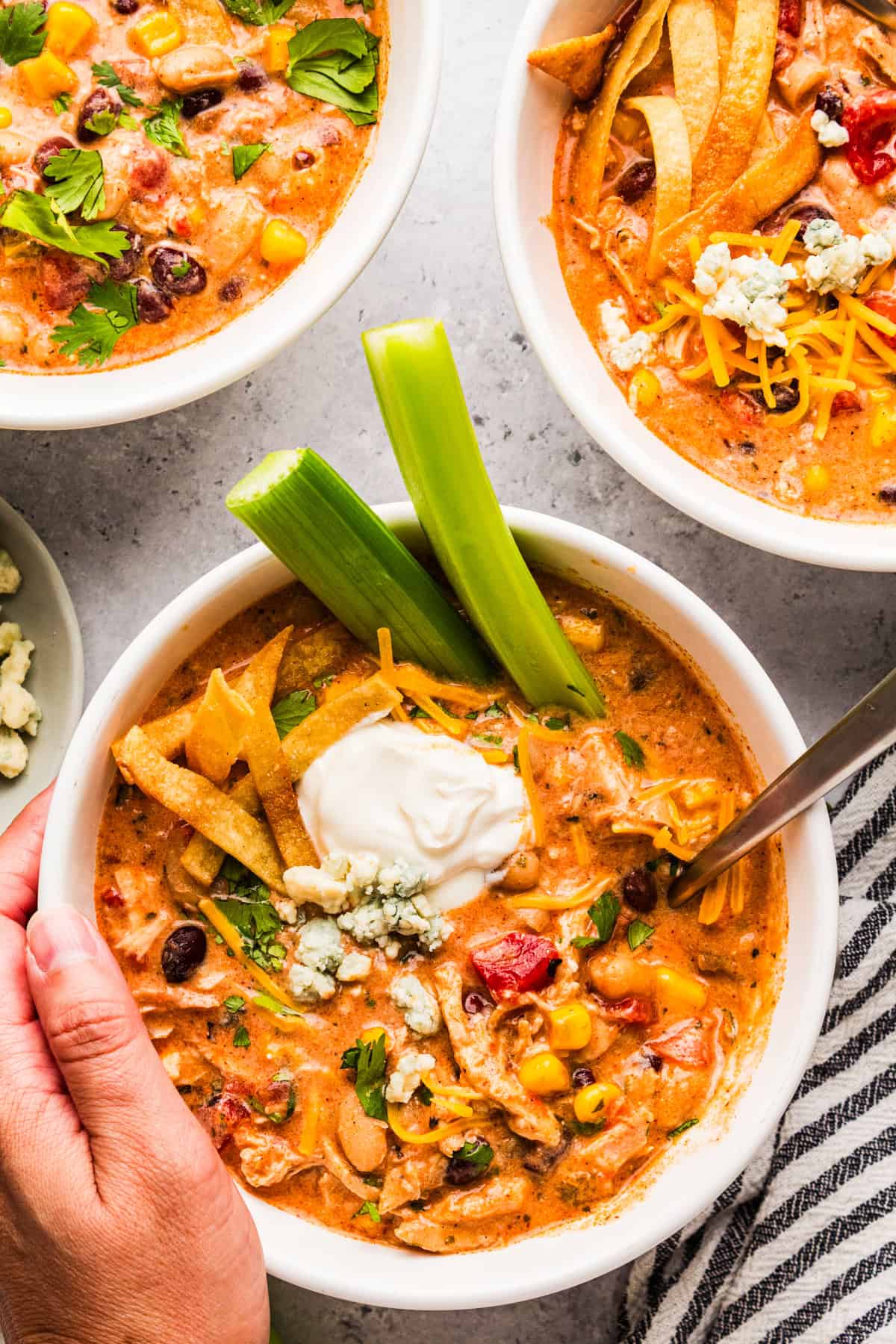 Thick and creamy Buffalo chicken chili prepared with shredded chicken, beans, Buffalo sauce, veggies, and cream cheese. An easy crockpot dinner for weeknights or game day! Bowls of Buffalo chicken chili topped with sour cream, blue cheese, cheddar, tortilla strips, and celery.