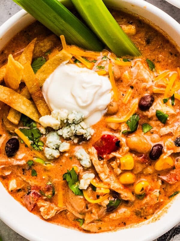 A serving of Buffalo chicken chili in a bowl, topped with a dollop of sour cream and shredded cheese, and two celery sticks.