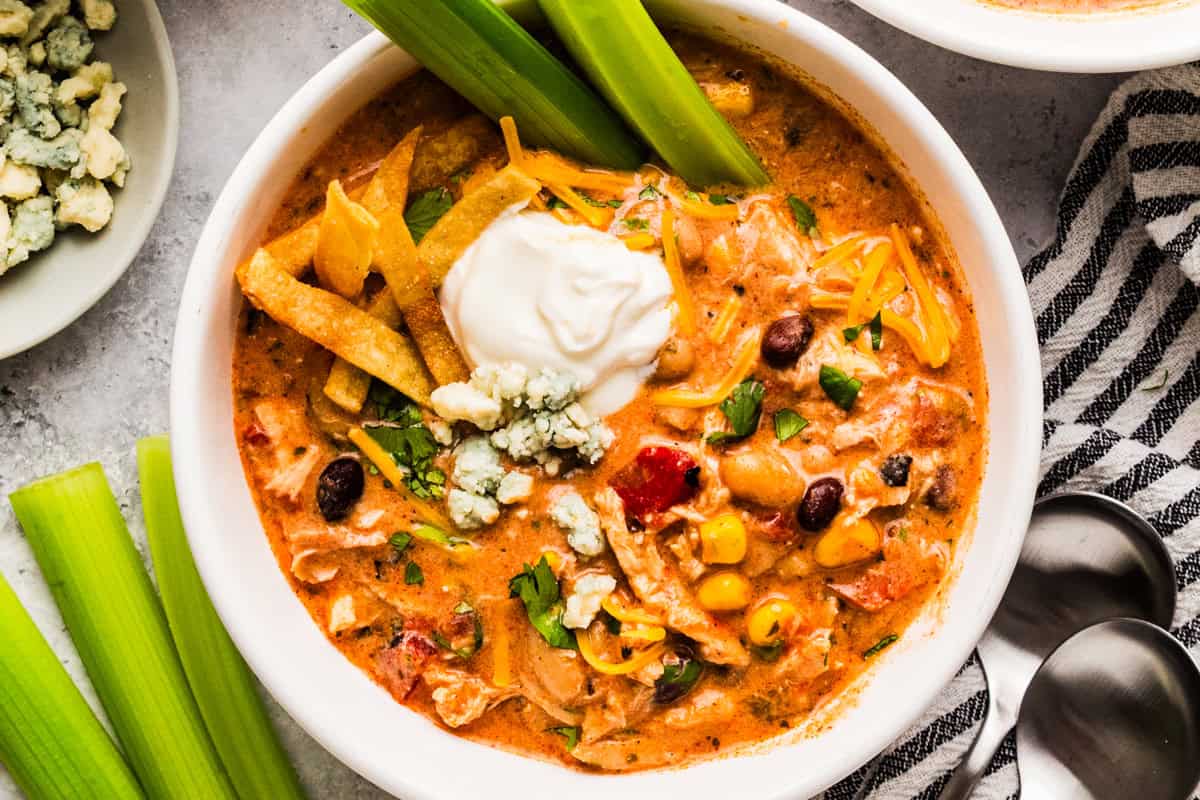 Thick and creamy Buffalo chicken chili prepared with shredded chicken, beans, Buffalo sauce, veggies, and cream cheese. An easy crockpot dinner for weeknights or game day! A serving of Buffalo chicken chili in a bowl, topped with a dollop of sour cream and shredded cheese, and two celery sticks.