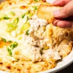 Close-up of a wonton chip lifting creamy, cheesy crab rangoon dip from a baking dish.